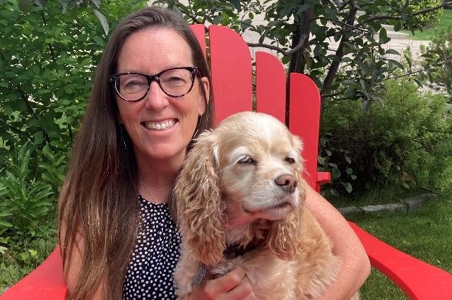 Rachel Syme sitting down on a big red chair with a small dog on her lap smiling